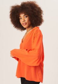 Loose-fitting, long-sleeve, orange blouse with a V-neckline. Smooth texture and slight draping. Accented with a subtle gathered detail at shoulders.