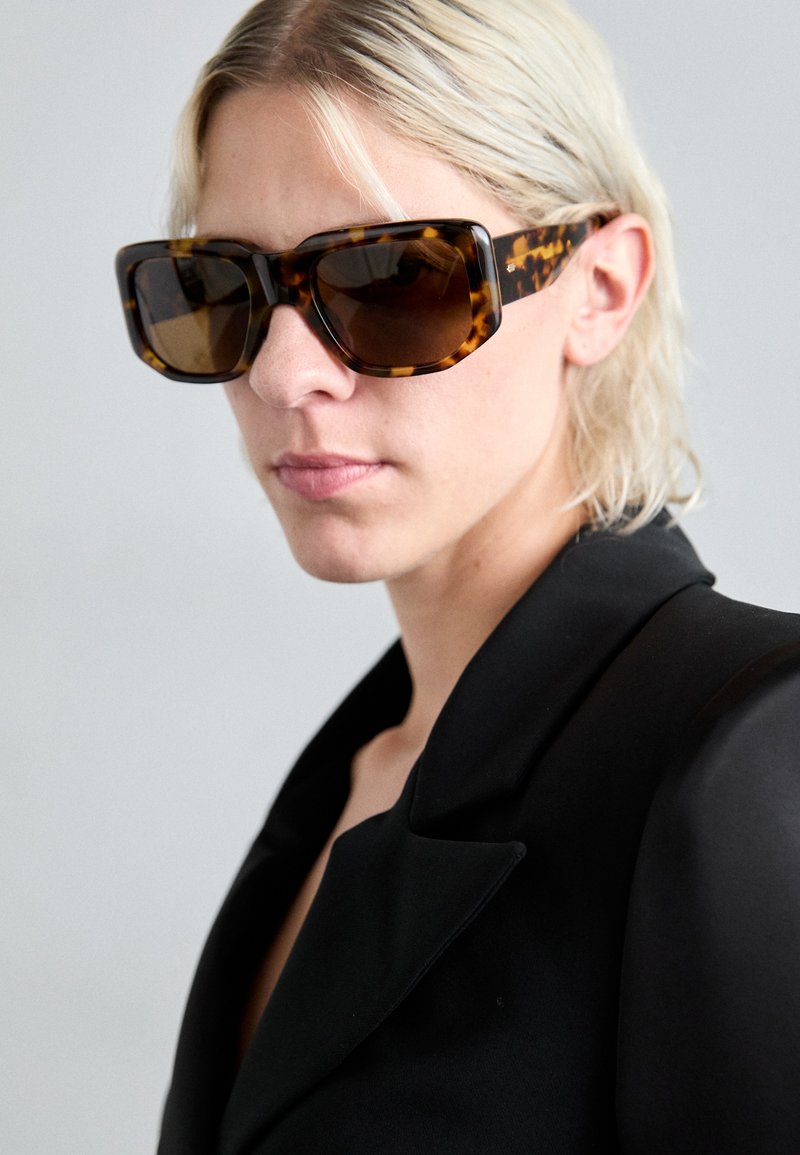 Lunettes de soleil surdimensionnées avec une monture écaille de tortue et des verres dégradés marron. Portées avec un blazer noir, mettant en avant un design angulaire et une texture brillante.