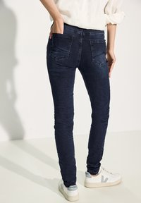 Jeans skinny bleu foncé avec une texture lisse, dotés de poches arrière et d'une taille mi-haute. Associés à des baskets blanches.