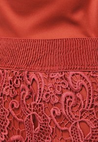 Tissu rouge avec une ceinture côtelée, orné de détails en dentelle complexe en bas, présentant des motifs floraux et des textures en relief.