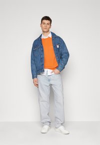 Denim jack met twee borstzakken, gecombineerd met een oranje trui, een wit overhemd met kraag, lichte spijkerbroek en witte sneakers.