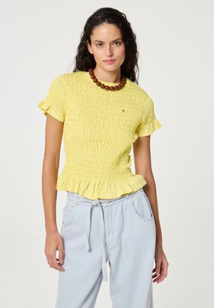 Woman with curly dark hair wears a yellow smocked top with ruffled edges and light blue drawstring pants, accessorized with a chunky brown necklace.