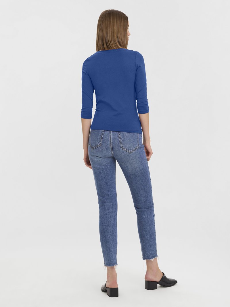 Blaues langärmliges Oberteil kombiniert mit hellblauen, eng anliegenden Jeans. Die Jeans haben ausgefranste Säume. Schwarze Blockabsatz-Mules vervollständigen das Outfit.