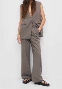 Grå skräddarsydda vida byxor som matchas med en ärmlös blazer. Tyget har en slät textur. Bärs med enkla svarta sandaler.