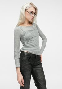 Šviesiaplaukė moteris, vilkinti pusperį pilką ilgomis rankovėmis marškinėlį, juodomis odinėmis kelnėmis ir skaidriais akinių rėmeliais, žiūri žemyn, ranka ant klubų.