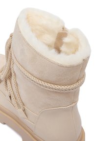 Cizmă de gleznă bej din întoarsă cu căptușeală din blană sintetică, șireturi împletite și o talpă groasă. Cizma are un detaliu din piele netedă.