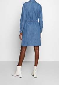 Denim-nappimekko, jossa pitkät hihat, vyötäröltä kiristetty leikku ja polven pituinen helma. Yhdistetty valkoisiin, korollisiin nilkkureihin. Pehmeä ja tekstuuri kangas.