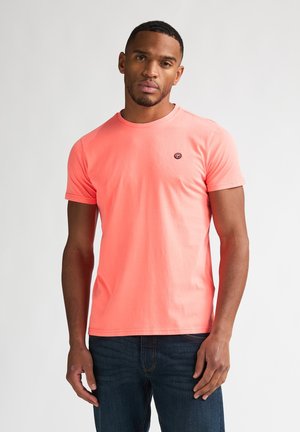 Man draagt een koraalkleurig T-shirt met korte mouwen en een klein logo op de borst en donkerblauwe spijkerbroek, staand tegen een witte achtergrond.