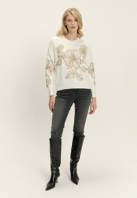 Eine Frau trägt einen weißen Pullover mit großen braunen Blumenmustern, dunkelgraue Jeans und schwarze kniehohe High Heels. Sie steht vor einem schlichten Hintergrund.