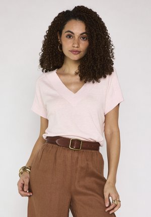 Donna con capelli ricci che indossa una maglietta rosa chiaro a V, pantaloni marroni con cintura, orecchini a cerchio dorati e braccialetti su uno sfondo uniforme.