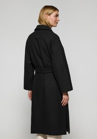 Abrigo de lana negro con cuello, mangas largas, cinturón y aberturas laterales. Textura suave y diseño minimalista. Vista trasera mostrada.