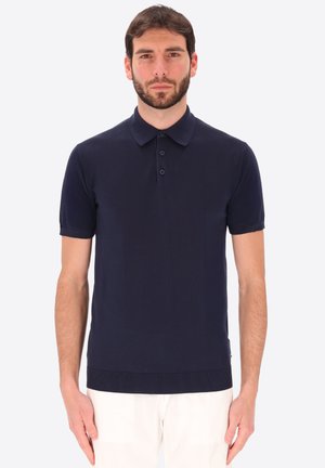 Uomo in piedi rivolto in avanti, che indossa una polo blu navy a maniche corte e pantaloni bianchi su uno sfondo semplice.