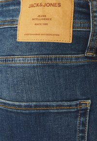 Mörkblå denimjeans med en synlig läderetikett, med sömnadsdetaljer och kontrasterande orange tråd på bakfickan.