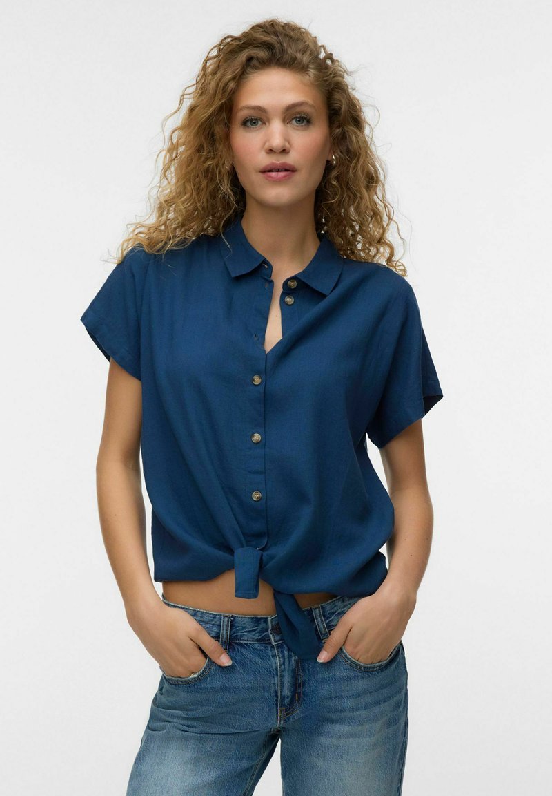 Femme aux cheveux bouclés portant une chemise bleu marine à manches courtes boutonnée et nouée à la taille, avec un jean bleu, mains dans les poches, fond blanc.