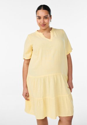 Kobieta z ciemnymi włosami związanymi do tyłu, nosząca złote kolczyki koła i luźną, krótką, jasnopomarańczową sukienkę, stojąca na białym tle.