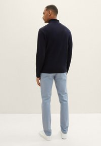 Man wearing a navy turtleneck sweater, light blue pants, and white sneakers standing facing away against a plain light background.