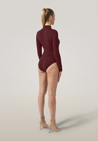 Kvinna med hästsvans som bär en långärmad bordeaux bodysuit och beige högklackade skor, stående med ryggen mot en enfärgad bakgrund.