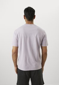 Lavendel Baumwoll-T-Shirt mit kurzen Ärmeln, Rundhalsausschnitt und einer Rückennaht-Detail. Getragen mit dunklen Shorts. Glatte Textur.