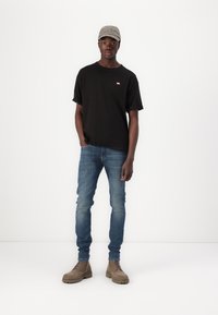 Svart t-skjorte med liten logo, blå denimjeans og beige snøresko. Bærer en grå cap med brodert tekst. Ensfarget hvit bakgrunn.