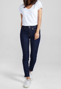 Jeans skinny de talle alto en azul oscuro con diseño de cinco bolsillos, costuras en contraste y cierre de botón metálico. Combinados con una camiseta blanca de cuello en V.