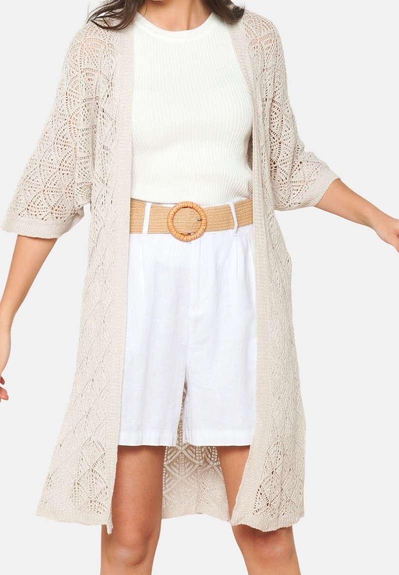 Cardigan en tricot beige avec une ouverture à l'avant, motif complexe et manches trois-quarts, porté sur un top blanc à côtes et un short ceinturé.