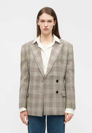 Γυναίκα που φοράει φαρδιά μπεζ καρό σακάκι διπλής σειράς κουμπιών πάνω από λευκό πουκάμισο και μπλε τζιν, στέκεται μπροστά σε απλό ανοιχτό φόντο.