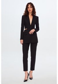 Svart skräddarsydd kostym med en kort blazer med djup V-ringning, två framfickor och smala byxor. Stylingen kompletteras med svarta högklackade skor.