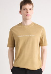 Lühikeste varrukatega, ümmarguse kaelusega t-särk heleses pruunis värvitoonis, millel on valge kiri "BARBOUR INTERNATIONAL" rinna peal. Pehme tekstuur.