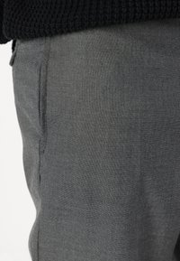Szürke szövetnadrág, texturált felülettel, oldalsó zsebbel és slim fit szabással, részben egy fekete kötött felső takarja.