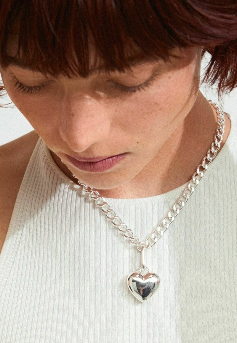 Collier en chaîne argentée avec un pendentif en forme de cœur poli, contre un haut à rayures légères, mettant en valeur la texture et l'éclat métallique.