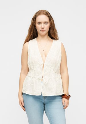 Kobieta o długich rudych włosach, ubrana w białą koronkową bluzkę na ramiączkach z wiązaniami z przodu, jasnoniebieskie dżinsy, naszyjnik i drewniane bransoletki na lewym nadgarstku.