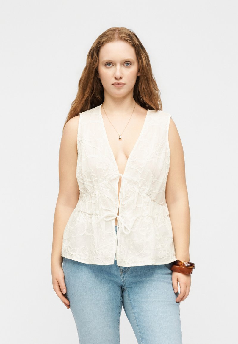 Γυναίκα με μακριά κόκκινα μαλλιά, που φοράει ένα λευκό αμάνικο δαντελένιο τοπ με κορδόνια μπροστά, ανοιχτό μπλε τζιν, ένα κολιέ και ξύλινες βραχιόλια στον αριστερό καρπό.