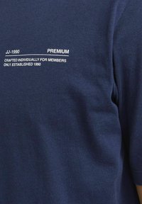 T-maica temno modre barve s kratkimi rokavi, ki vsebuje belo besedilo: "JJ-1990", "PREMIUM" in "ROČNO IZDELANO ZA ČLANE SAMO. USTANOVLJENO 1990."