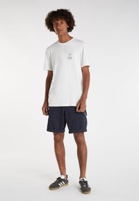 Witte katoenen t-shirt met ronde hals, korte mouwen en een kleine borstafbeelding; gecombineerd met marineblauwe cargoshorts en grijze sneakers met witte strepen.