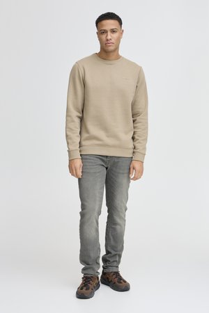 Sweatshirt beige à col rond, avec des poignets côtelés et un petit logo sur la poitrine, associé à un jean gris et des baskets marron.