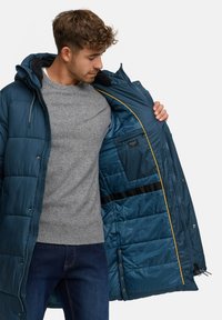 Jachetă puf albastră cu glugă căptușită, fermoare și detalii galbene. Strat interior cu un buzunar și textură matlasată. Pulover gri dedesubt.