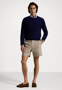 Granatowy sweter z grubym splotem, kremowe spodenki, brązowe mokasyny. Model ma na sobie pasiastą koszulę pod swetrem. Prosty, codzienny zestaw.