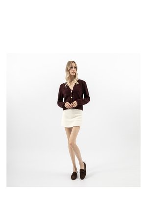 Frau trägt einen dunkelburgunderfarbenen, geknöpften Cardigan, einen weißen, strukturierten Minirock und dunkelbraune Loafers, steht vor weißem Hintergrund.