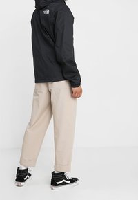 Černá voděodolná bunda s kapucí, s logem na zádech, v kombinaci s béžovými širokými kalhotami a černými teniskami.