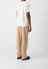 Bílý bavlněný tričko s kulatým výstřihem, kombinované s béžovými širokými kalhotami. Model má na sobě černou obuv, pohled zezadu.