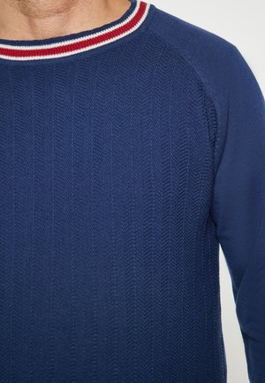 Blauwe gebreide trui met een gestructureerd haringbeenpatroon. Heeft een ronde halslijn met rode en witte gestreepte accenten.