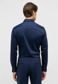 Man met kort bruin haar, gekleed in een nauwsluitend marineblauw overhemd en bijpassende marineblauwe broek, gezien van achter tegen een effen achtergrond.