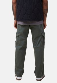 Tummansininen tikattu liivi harmaan paidan päällä, yhdistettynä löysiin oliivinvihreisiin cargo-housuihin, joissa on sivutaskut ja rento leikkaus. Ruskea-valkoiset lenkkarit.