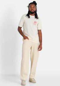 Lichtbeige T-shirt met korte mouwen met een roze bloemenafbeelding op de borst, gecombineerd met bijpassende beige wijde broek en off-white schoenen.