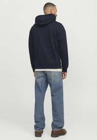 Haut à capuche bleu marine avec une finition texturée, doté d'une grande capuche et d'un ourlet côtelé, associé à un jean droit bleu clair et des chaussures foncées.