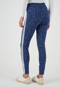 Donna che indossa leggings blu con stampa leopardata, strisce bianche ai lati e sneakers bianche con dettagli dorati, in piedi su uno sfondo bianco neutro.