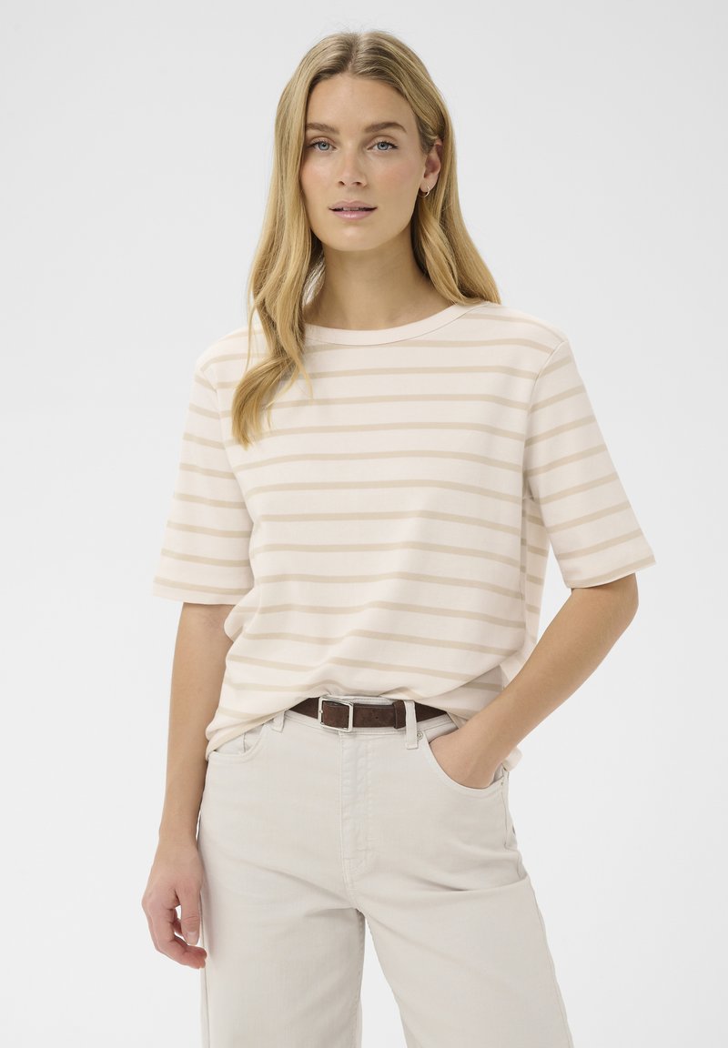 Femeie cu păr lung și blond, purtând un tricou beige cu dungi albe și mâneci scurte și pantaloni deschiși la culoare cu o centură închisă, stând în picioare.