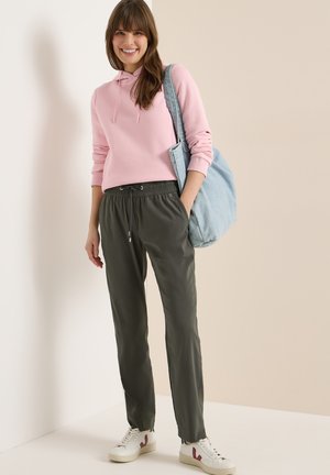 Jeune femme souriante, portant un sweat à capuche rose, un pantalon vert foncé, des baskets blanches, et portant un grand sac à bandoulière bleu clair sur un fond uni.