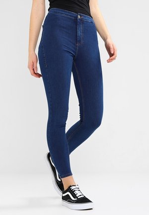Jeans ajustados de mezclilla en un tono oscuro, con una cintura alta, cierre frontal de botón y costuras en contraste. Llevados con zapatillas en blanco y negro.