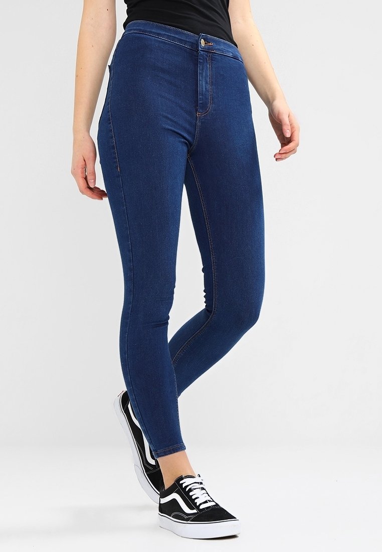 Jeans ajustados de mezclilla en un tono oscuro, con una cintura alta, cierre frontal de botón y costuras en contraste. Llevados con zapatillas en blanco y negro.